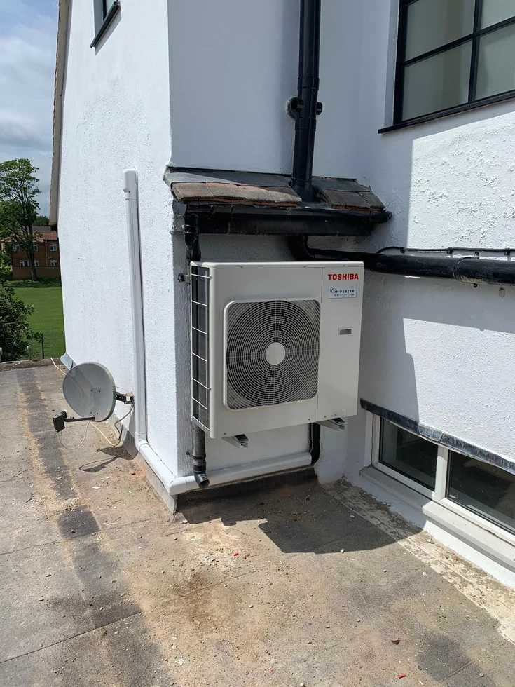 An outdoor air conditioning unit and pipework beside a white exterior wall.